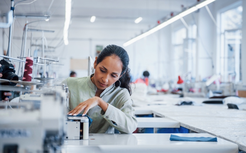 Woman wearing a green shirt with long hair sitting behind an industrial sewing machine inside a manufacturing sewing room.