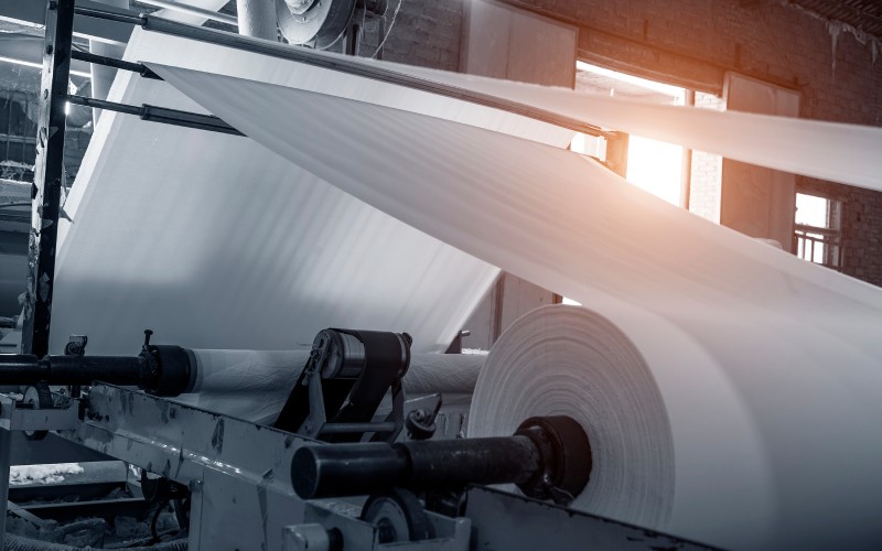Textile manufacturing machine producing a large roll of white fabric stretched through various tubes inside an industrial environment