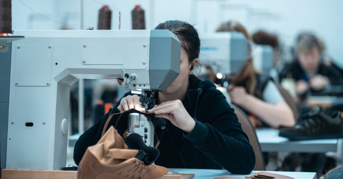 Multiple people sitting behind industrial sewing machines aligned in a vertical row inside a room with white walls.